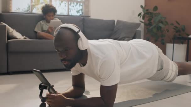 Foto completa de padre en auriculares manteniéndose en posición de tablón, siguiendo un tutorial de yoga en su teléfono mientras su hijo ve videos en una tableta digital - Metraje, vídeo