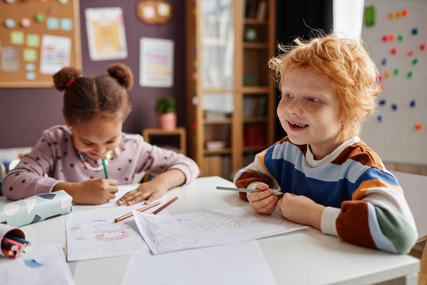Wesoły uroczy uczeń przedszkola z piegami i rudymi włosami trzymając ołówek siedząc przy stole przeciwko koledze z klasy i rysunek - Zdjęcie, obraz