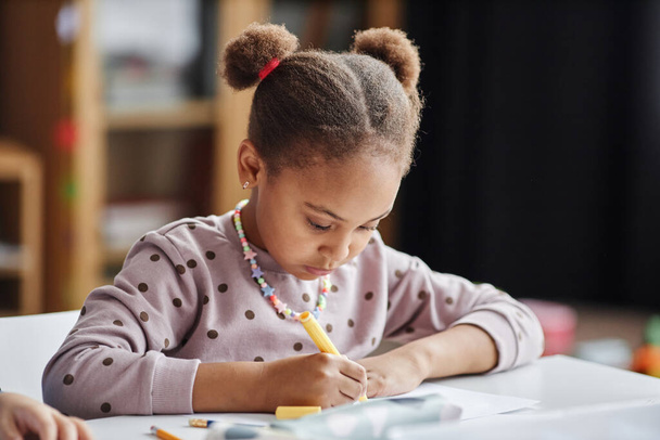 Cute poważne African American uczennica rysunek z żółtym podkreśleniem siedząc przy stole na lekcji rzemiosła i sztuki w przedszkolu - Zdjęcie, obraz