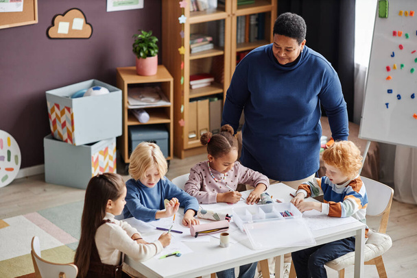 Grupa adorable uczących się przedszkola siedzi przy stole na lekcji, podczas gdy dojrzała Afroamerykanka nauczyciel stoi przy nich - Zdjęcie, obraz
