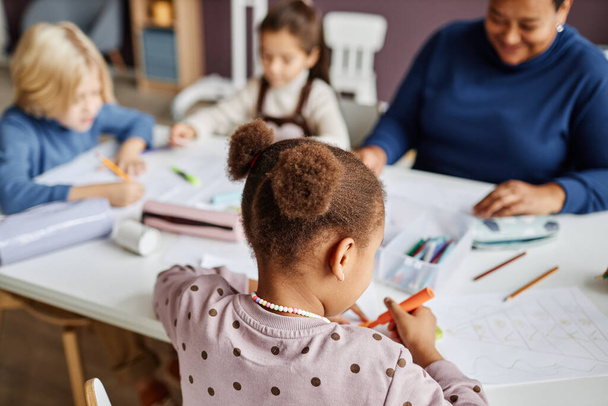 Tylny widok cute African American ucznia przedszkola rysunek podczas siedzenia przy stole przed dwoma kolegami z klasy i ich nauczyciel - Zdjęcie, obraz