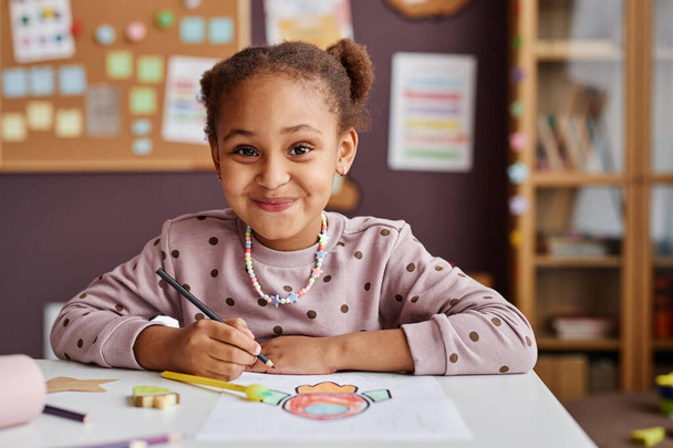 Roztomilý úspěšný žák základní školy při pohledu na fotoaparát s úsměvem při sezení u stolu a kreslení s pastelkami na lekci - Fotografie, Obrázek