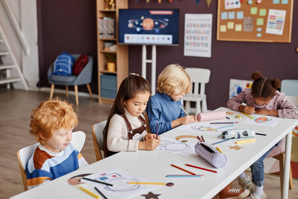 Varios lindos estudiantes interculturales de la escuela primaria sentados a la mesa en la lección en el jardín de infantes y dibujo con lápices de colores en los papeles - Foto, Imagen
