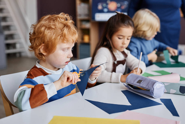 Cute pilny uczeń z nożyczkami wycinanie otworów na oczy w kolorze ciemnoniebieskim, siedząc przy biurku przed kolegami z klasy - Zdjęcie, obraz
