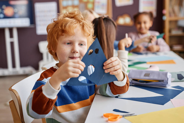 Netter Grundschüler hält blaues Papier mit Löchern für Augen und Mund, während er eine Gesichtsmaske für die Ferienaktion im Kindergarten kreiert - Foto, Bild