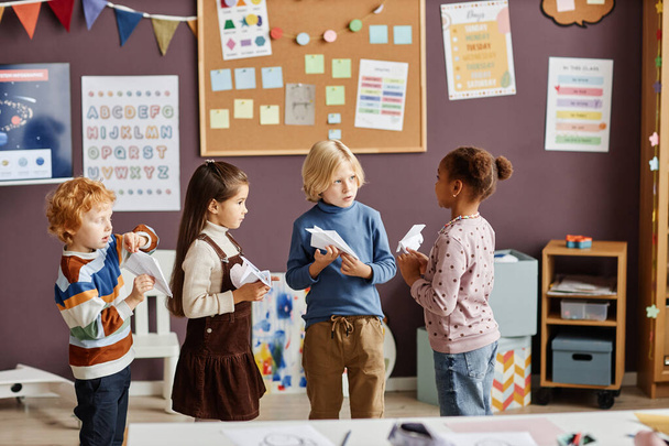 Группа из четырех межкультурных маленьких учащихся детского сада в повседневной одежде играет бумажные самолеты и взаимодействует во время перерыва в классе - Фото, изображение