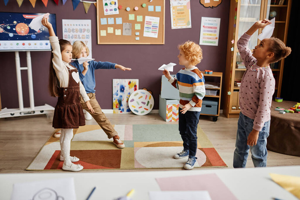 Група мультикультурних маленьких дітей грають у паперові літаки ручної роботи і розважаються між уроками в сучасній дитячій школі - Фото, зображення