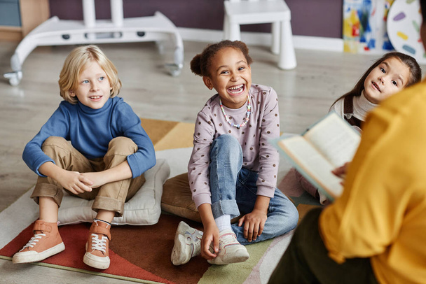 Skupina roztomilých malých žáků školky sedí na podlaze učebny a poslouchá učitele, jak si je čte. - Fotografie, Obrázek