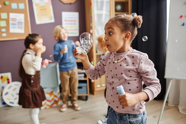 Sorprendida niña afroamericana mirando la burbuja de jabón múltiple mientras está de pie frente a la cámara contra sus compañeros de clase divirtiéndose - Foto, Imagen