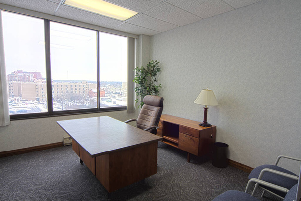 Un bureau exécutif professionnel à Fort Wayne, Indiana, doté d'un grand bureau en bois et d'une chaise en cuir à dossier haut, parfait pour un environnement de travail productif. La lumière naturelle et une vue urbaine ajoutent à - Photo, image