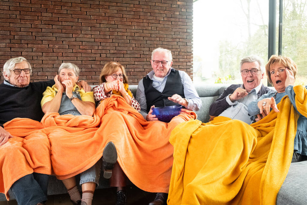 Cette image chaleureuse capture un groupe d'amis âgés partageant un moment de joie et de rire. Ils sont confortablement nichés sous une grande couverture orange vibrante, avec des expressions de bonheur authentique et - Photo, image