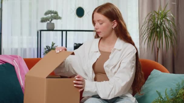 Onnellinen teini tyttö purkaa toimitus paketti istuu kotona huoneisto sohvalla. Hymyilevä lapsi shopper verkkokaupan asiakas avaa pahvilaatikon, joka vastaanottaa ostolahjan nopealta postilähetykseltä - Materiaali, video