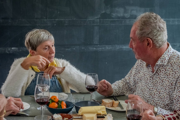 In questa suggestiva immagine, una coppia di anziani gode di un momento romantico, condividendo un brindisi con bicchieri di vino rosso. La tavola è allestita intimamente con un assortimento di formaggi e frutta fresca, a significare un - Foto, immagini