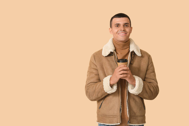 Young man in sheepskin jacket with cup of coffee on beige background - Photo, Image