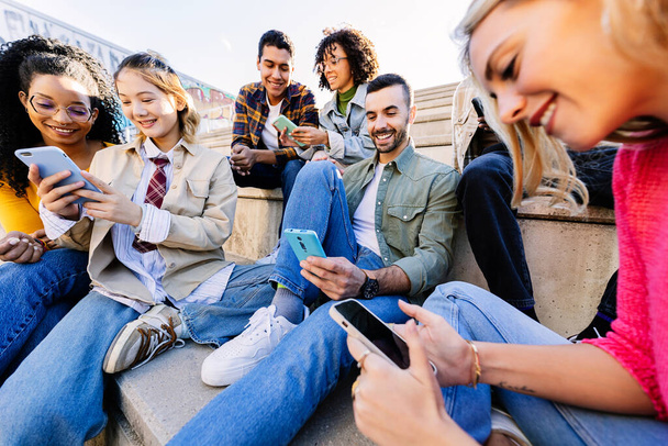 Залежні молоді люди використовують мобільні телефони, сидячи разом на відкритому повітрі. Тисячолітню групу різноманітних друзів, які дивляться на екран мобільного телефону, грають у відеоігри або насолоджуються вмістом соціальних мереж у додатку. - Фото, зображення