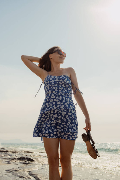 Çiçekli mavi elbiseli güzel bir esmer kadın kayalık bir plajda yürüyor, elinde bir çift sandaletle. - Fotoğraf, Görsel