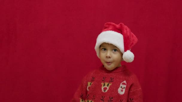 Lindo niño preescolar en el saltador de Navidad ondeando signo de Santa parada aquí mientras se come sus galletas. Movimiento lento aislado en rojo. Imágenes de alta calidad 4k - Metraje, vídeo