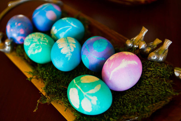 鮮やかなイースターエッグは,青と紫の色合いで染められ,葉のパターンで飾られ,緑色のモスで素朴な木製のトレイに設定されています. 小さな鳥のフィギュアは春の喜びのタッチを追加します. 伝統を祝う. - 写真・画像