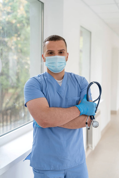 doutor masculino caucasiano em um casaco azul e uma máscara médica no corredor da clínica. Local de trabalho - Foto, Imagem