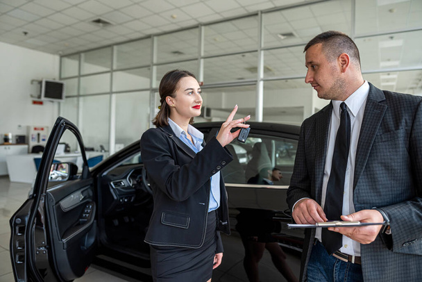  Όμορφη νεαρή γυναίκα στέκεται κοντά στο νέο σύγχρονο αυτοκίνητο στην αντιπροσωπεία και επιλέγοντας το καλύτερο αυτοκίνητο με τη βοήθεια του πωλητή. Έννοια της αγοράς - Φωτογραφία, εικόνα