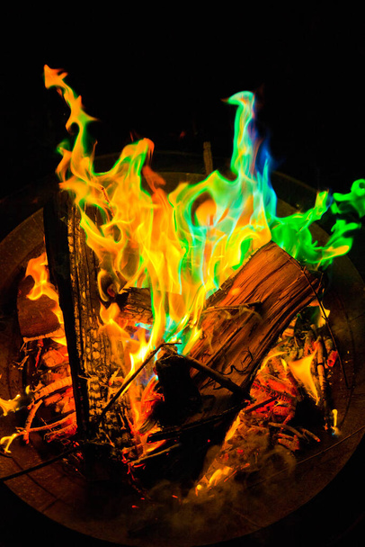 Sperimenta il fascino ipnotizzante di un fuoco vibrante e multicolore in questa accattivante foto stock. Immerse in una fossa di fuoco metallica, le fiamme danzano con toni verdi, blu e tradizionali giallo-arancio. - Foto, immagini