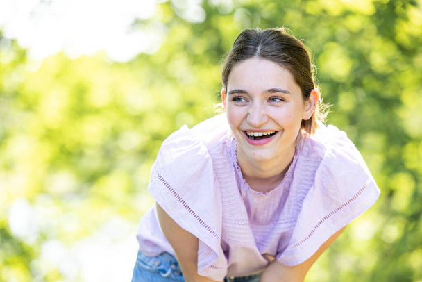 A joyful young woman leans forward with a candid laugh, her eyes sparkling with mirth. She is outdoors, surrounded by the bright green hues of a park in full bloom. The blurred background suggests a - Photo, Image