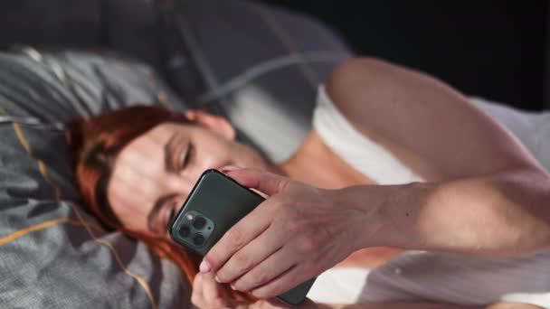 jovem mulher usando telefone celular e mensagens de texto com amigos enquanto deitado na cama no dia ensolarado - Filmagem, Vídeo