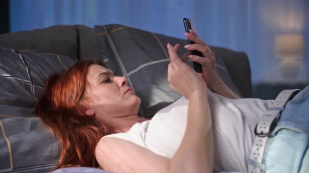 мила молода жінка використовує мобільний телефон і читає новини в Інтернеті, лежачи на дивані вдома - Кадри, відео