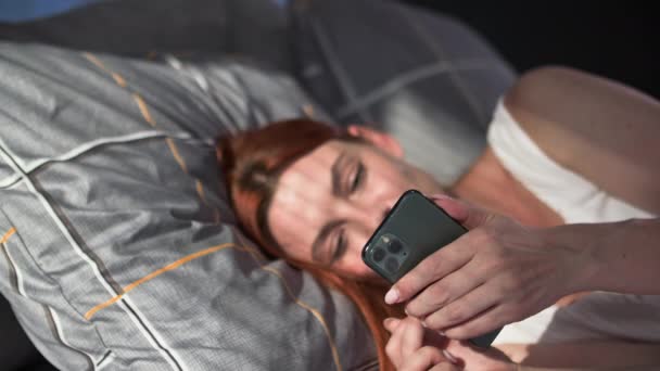nainen käyttää matkapuhelinta kommunikoida ystävien kanssa sosiaalisissa verkostoissa tai tehdä ostoksia verkossa rentoutuessaan makaamassa sohvalla kotona - Materiaali, video