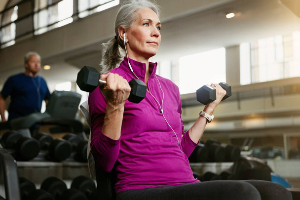 Idős nő, fitness és súlyzó edzőteremben edzés, wellness és kardió fülhallgatóval, zene vagy gondolkodásmód. Súlyemelés, testépítés és idősebb női személy egy sportközpontban bicepsz edzés. - Fotó, kép