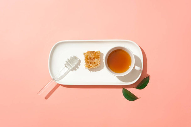 Ελάχιστη σκηνή μελιού που στάζει, κερί μέλισσας και ένα φλιτζάνι τσάι τοποθετημένα σε μια γραμμή σε ένα πιάτο. Ροζ φόντο. Το μέλι εξακολουθεί να περιέχει υψηλές θερμίδες και ζάχαρη - Φωτογραφία, εικόνα