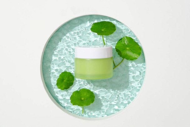 Een pot groene lotion en pennywort bladeren worden geplaatst op een glazen platform met een rimpel effect. Blanco label voor belettering ontwerp. Ruimte voor reclame. - Foto, afbeelding
