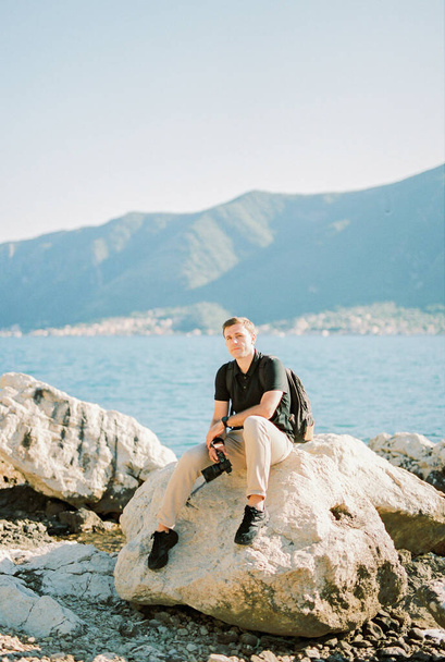 Photographe avec un appareil photo à la main assis sur un grand rocher au bord de la mer. Photo de haute qualité - Photo, image