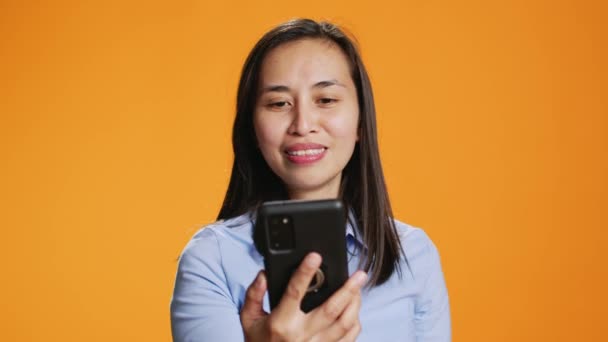 Mujer asiática responde videocall en línea en el estudio, hablando con la gente en la conexión remota a Internet. Joven adulto sonriendo y saludando en la pantalla del teléfono, reunión de videoconferencia en línea sobre el fondo. - Metraje, vídeo