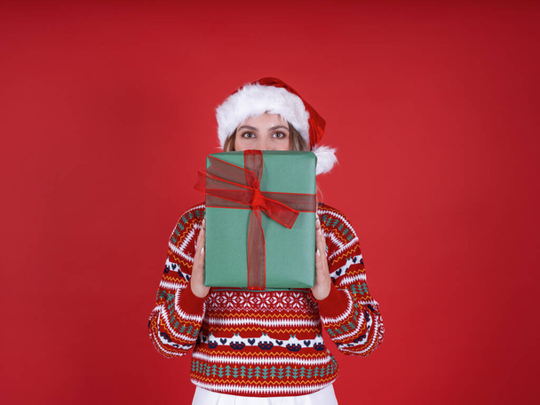 Žena si zakrývající obličej dárkovou krabičkou, portrét krásné bílé blondýnky, která si zakrývá obličej dárkovou krabičkou. Usmívající se dívka vrcholí nad přítomností. Noste Santa klobouk, vánoční svetr. - Fotografie, Obrázek