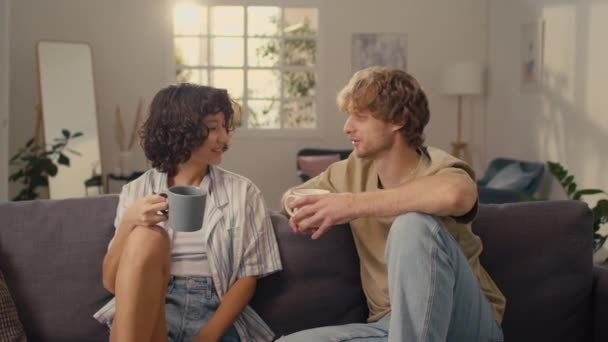 Mediana toma de pareja de jóvenes casados sentados en el sofá, tomando bebidas calientes y hablando entre sí - Imágenes, Vídeo