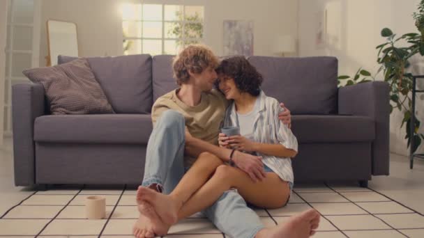 Foto completa de pareja joven sentados juntos en el suelo de su espaciosa sala de estar mientras la mujer sostiene la taza de bebida caliente - Imágenes, Vídeo