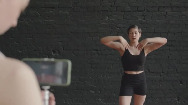 黒いアクティブウェアで若いエレガントな白人女性バレエダンサーの肩のショットに黒いレンガの背景でバレエを踊る彼女の学生がスマートフォンでそれのビデオを録画しながら - 映像、動画