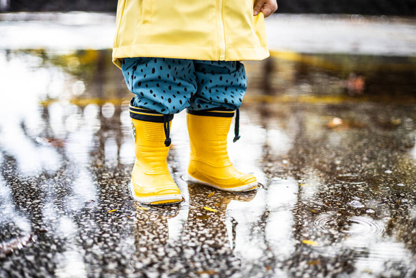 Piccolo bambino che indossa stivali di gomma gialli e impermeabile giallo in piedi in pozzanghera in un giorno di pioggia nuvoloso. Bambino sotto la pioggia - Foto, immagini
