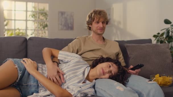 Medium laukaus nuori mies vaaleat hiukset istuu sohvalla, kun hänen tyttöystävänsä makaa sylissään iloisesti keskustella elokuvan he katsovat - Materiaali, video