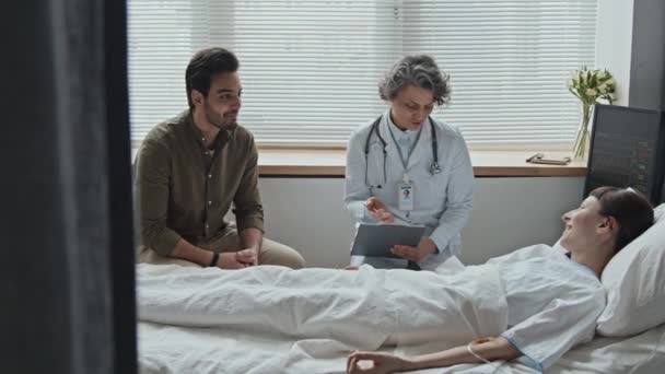 Female doctor talking with interracial couple about prospects of recovery and necessary treatment for woman lying in bed while her Middle Eastern boyfriend attentively listening to specialist - Footage, Video