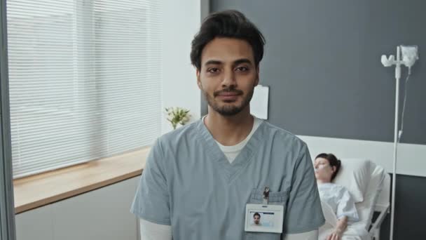 Muotokuva Lähi-idän miespuolisesta sairaanhoitajasta, joka seisoo sairaalaosastolla lääketieteellisissä vaatteissa katsoen kameraa, kun hänen potilaansa makaa sängyssä taustalla - Materiaali, video