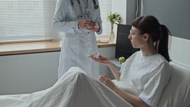 Keskisuuren kuvan lääkäri tuo lääkettä ja hänen naispuolinen potilas ottaa pillereitä istuessaan sairaalassa sängyssä - Materiaali, video