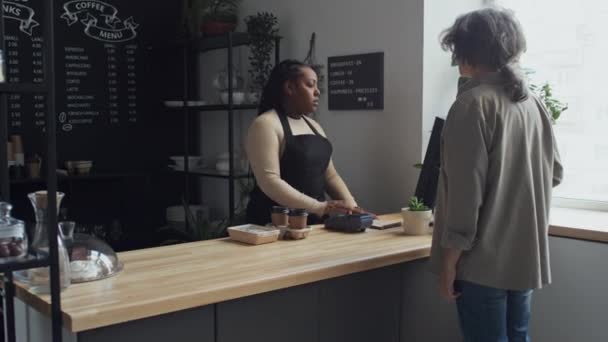 Medium long shot of female customer with grey hair paying for her order with smartphone through terminal with barista standing behind counter in coffee shop - Footage, Video