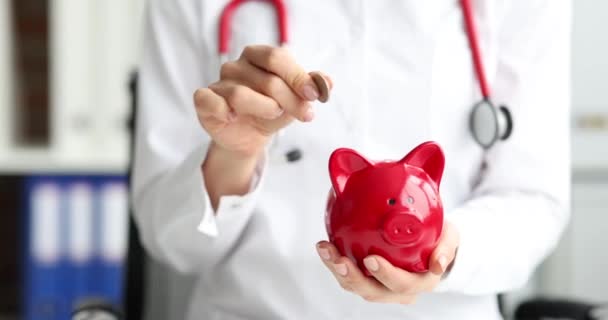 Doctor tosses coin with red pig to piggy bank. Life and health insurance concept - Footage, Video