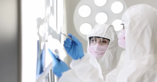 Δύο γιατροί με προστατευτικές στολές και μάσκες εξετάζουν ακτινογραφίες στο εργαστήριο. Έννοια διαγνωστικού εξοπλισμού εστίας του Coronavirus - Πλάνα, βίντεο