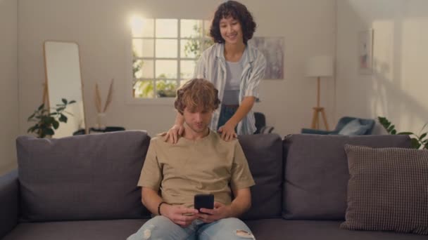 Giovane uomo con i capelli biondi seduto sul divano guardando attraverso il suo telefono mentre sua moglie viene in soggiorno e parla con lui - Filmati, video