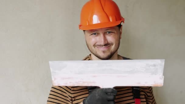 Glimlachende klusjesman reparateur in overalls en beschermende oranje helm houdt grote stopverf mes voor het pleisteren van betonnen muur met stopverf. Renovatie appartement en nieuwbouw concept - Video
