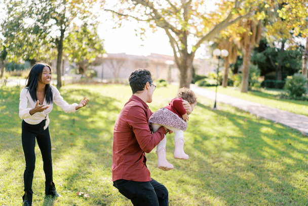 Η γελαστή μαμά απλώνει τα χέρια της στο κοριτσάκι που περικυκλώνει τον μπαμπά στο πάρκο. Υψηλής ποιότητας φωτογραφία - Φωτογραφία, εικόνα