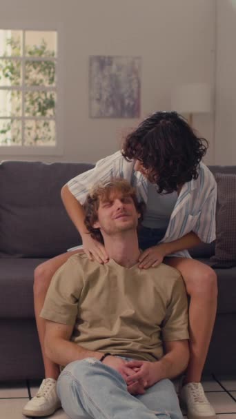 Κάθετη μέτρια βολή της νεαρής γυναίκας μασάζ ώμους του συζύγου της, ενώ αυτός κάθεται στο πάτωμα χαλαρώνοντας - Πλάνα, βίντεο